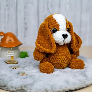 May include: A handmade, plush dog toy in shades of brown and white. The stuffed animal has floppy ears, a white patch on its head, and a black nose. It is sitting on a gray, fluffy surface.
