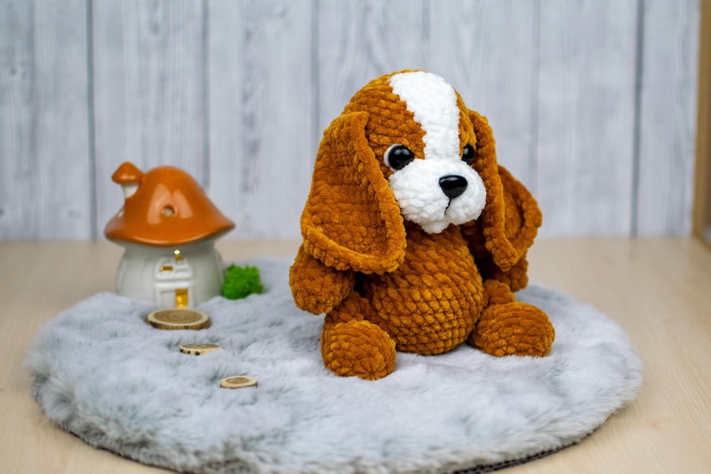 May include: A handmade, plush dog toy in shades of brown and white. The stuffed animal has floppy ears, a white patch on its head, and a black nose. It is sitting on a gray, fluffy surface.