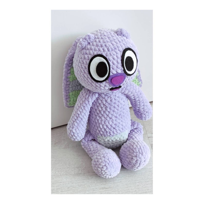 May include: A plush toy crafted from light purple textured yarn, featuring long ears with light green accents, large white eyes with black pupils, a purple nose, and a small mouth. The stuffed animal has a light green patch on its belly.
