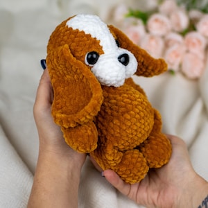 May include: A handmade, plush dog toy in shades of brown, white, and black. The crochet dog has large, floppy ears and a soft, textured appearance. The toy is held in a person's hands.