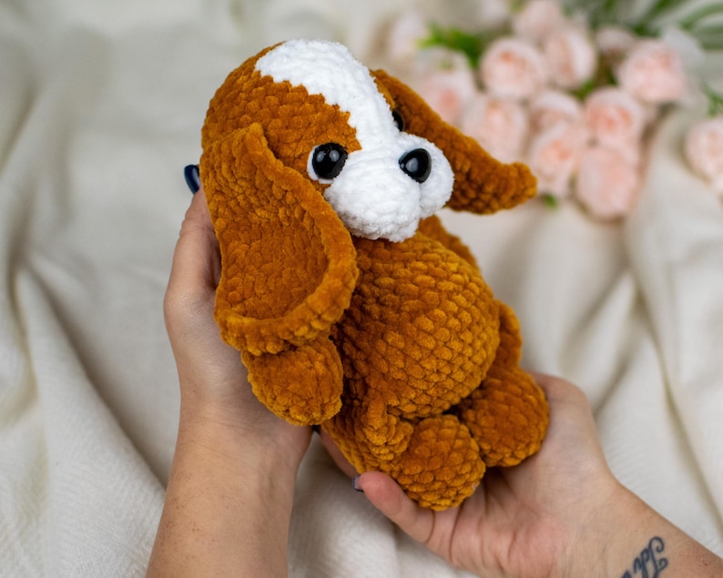 May include: A handmade, plush dog toy in shades of brown, white, and black. The crochet dog has large, floppy ears and a soft, textured appearance. The toy is held in a person's hands.