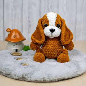May include: A handmade, plush dog toy in shades of brown and white, with large, floppy ears and a sweet expression. The dog is sitting on a gray, round rug. A small ceramic house with an orange roof is in the background.