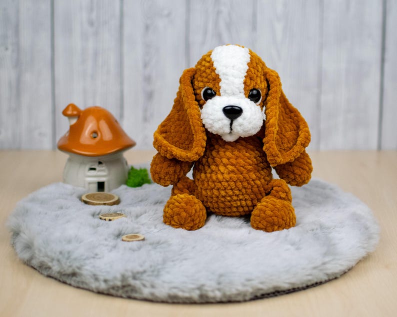 May include: A handmade, plush dog toy in shades of brown and white, with large, floppy ears and a sweet expression. The dog is sitting on a gray, round rug. A small ceramic house with an orange roof is in the background.