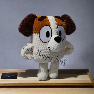 May include: A crocheted plush toy dog with brown ears, white fur, and brown spots. The dog has large white eyes and a black nose. The toy is standing on a wooden surface.