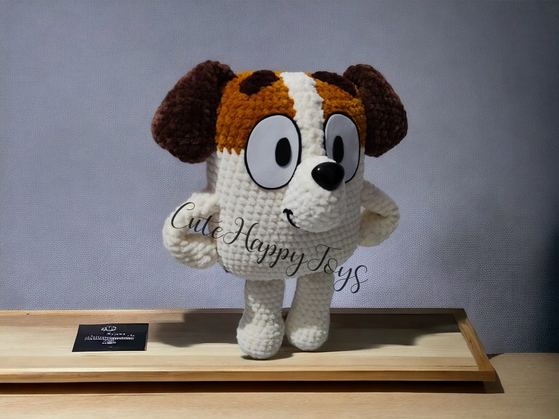 May include: A crocheted plush toy dog with brown ears, white fur, and brown spots. The dog has large white eyes and a black nose. The toy is standing on a wooden surface.