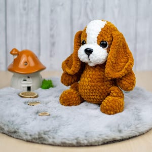 May include: A handmade, plush dog toy with brown and white fur, large floppy ears, and big black eyes. The stuffed animal is sitting on a gray, fluffy surface. A small ceramic mushroom house is in the background.