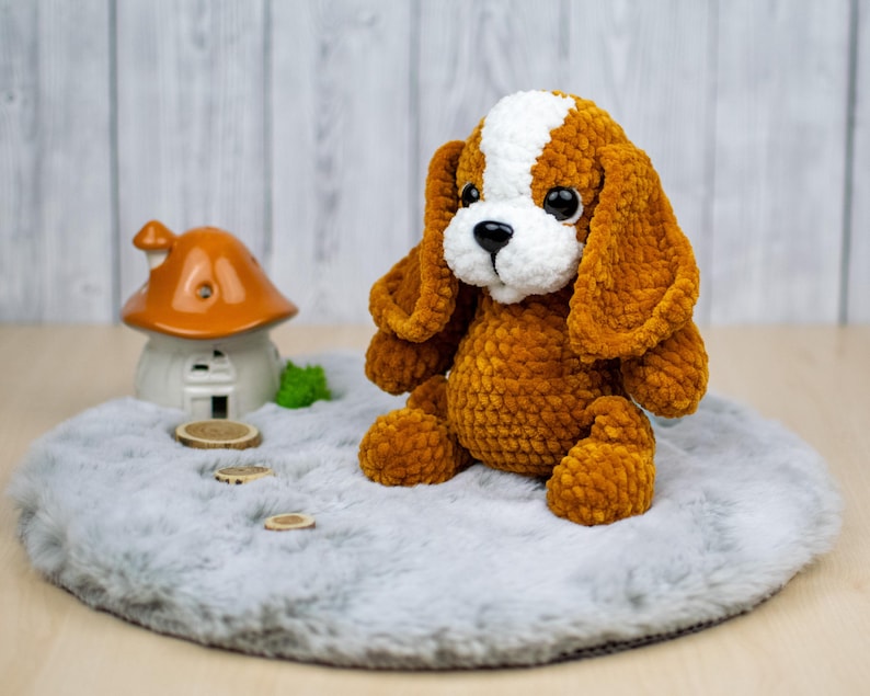 May include: A handmade, plush dog toy with brown and white fur, large floppy ears, and big black eyes. The stuffed animal is sitting on a gray, fluffy surface. A small ceramic mushroom house is in the background.