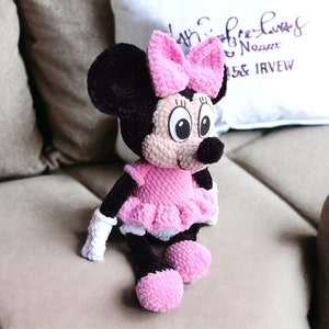 May include: A handmade Minnie Mouse plush doll. The doll features a black head with large, round eyes, a pink bow, and a black nose. It wears a pink dress with a ruffled skirt, white gloves, and pink shoes. The doll is sitting on a beige couch.