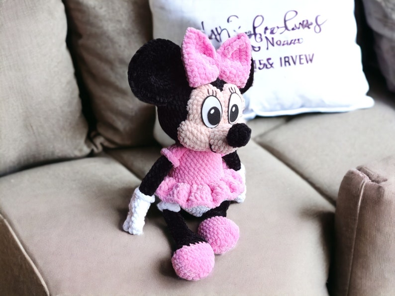 May include: A handmade Minnie Mouse plush doll. The doll features a black head with large, round eyes, a pink bow, and a black nose. It wears a pink dress with a ruffled skirt, white gloves, and pink shoes. The doll is sitting on a beige couch.