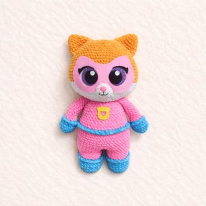May include: A handmade crochet superhero cat doll. The doll has an orange head with cat ears and a pink mask. It wears a pink suit with blue accents and a yellow emblem. The eyes are large and purple.
