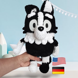 Può includere: Peluche di cane in crochet nero e bianco con occhi grandi e un colletto bianco. Il giocattolo è tenuto nella mano di una persona. Lo sfondo è una parete blu chiaro con pompon bianchi.