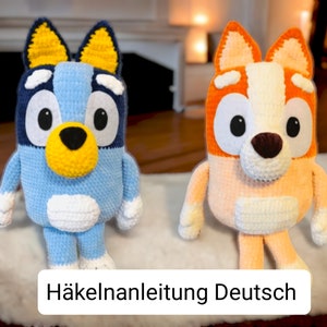 May include: Two crocheted stuffed dog toys, one blue and one orange, with white accents. The toys are sitting on a white fluffy surface. The text "H&auml;kelanleitung Deutsch" is visible at the bottom of the image.