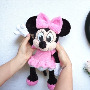 May include: A handmade crochet Minnie Mouse doll. The doll features a pink dress and bow, black ears, and white gloves. The doll is held up against a white wall.