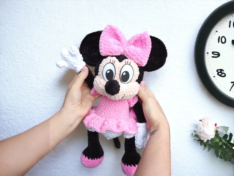 May include: A handmade crochet Minnie Mouse doll. The doll features a pink dress and bow, black ears, and white gloves. The doll is held up against a white wall.