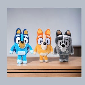 May include: Three handmade crochet dog figures in blue, orange, and grey, each with large eyes and a distinctive snout. The figures are standing on a wooden surface, showcasing the craftsmanship of the plush toys.