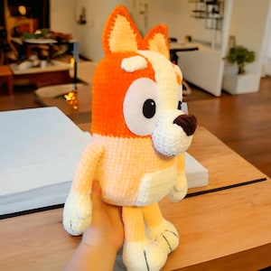 May include: A crocheted plush toy dog with orange and white fur. The dog has large, black eyes and a brown nose. The toy is sitting on a wooden table.