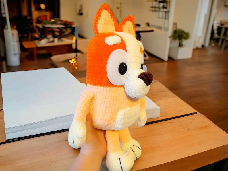 May include: A crocheted plush toy dog with orange and white fur. The dog has large, black eyes and a brown nose. The toy is sitting on a wooden table.