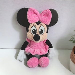 May include: A plush Minnie Mouse doll, featuring a black head with large ears and a pink bow. The doll has a pink dress, matching shoes, and white gloves. The doll is made of a soft, textured material.