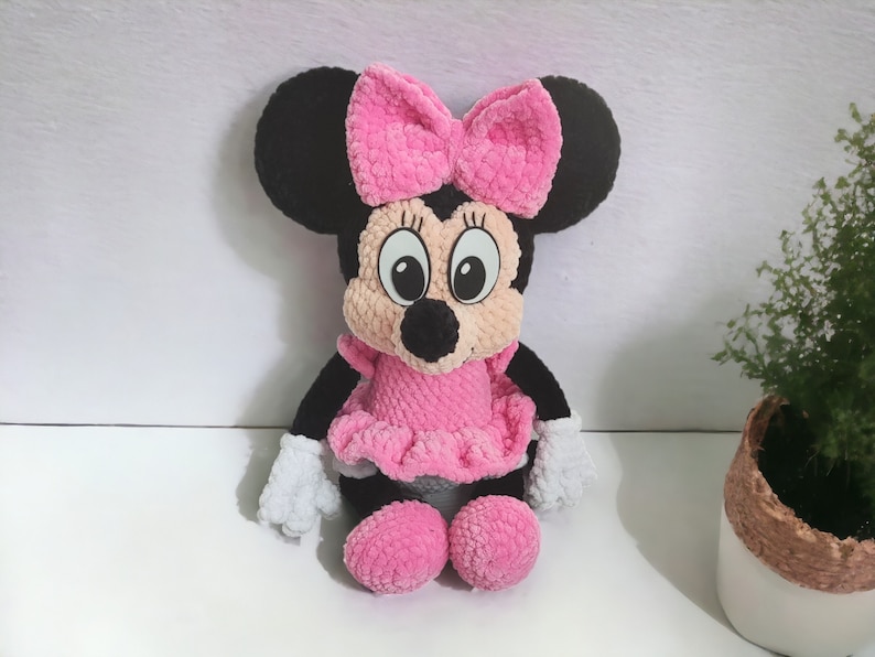 May include: A plush Minnie Mouse doll, featuring a black head with large ears and a pink bow. The doll has a pink dress, matching shoes, and white gloves. The doll is made of a soft, textured material.