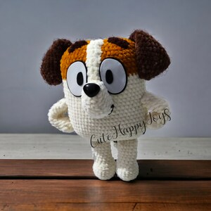 May include: A crocheted stuffed dog toy with brown and white fur, large eyes, and a black nose. The toy is standing on a wooden surface and has the text "Cute Happy Toys" on its side.