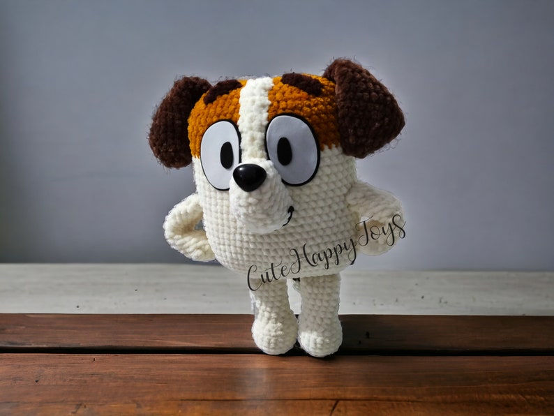 May include: A crocheted stuffed dog toy with brown and white fur, large eyes, and a black nose. The toy is standing on a wooden surface and has the text "Cute Happy Toys" on its side.