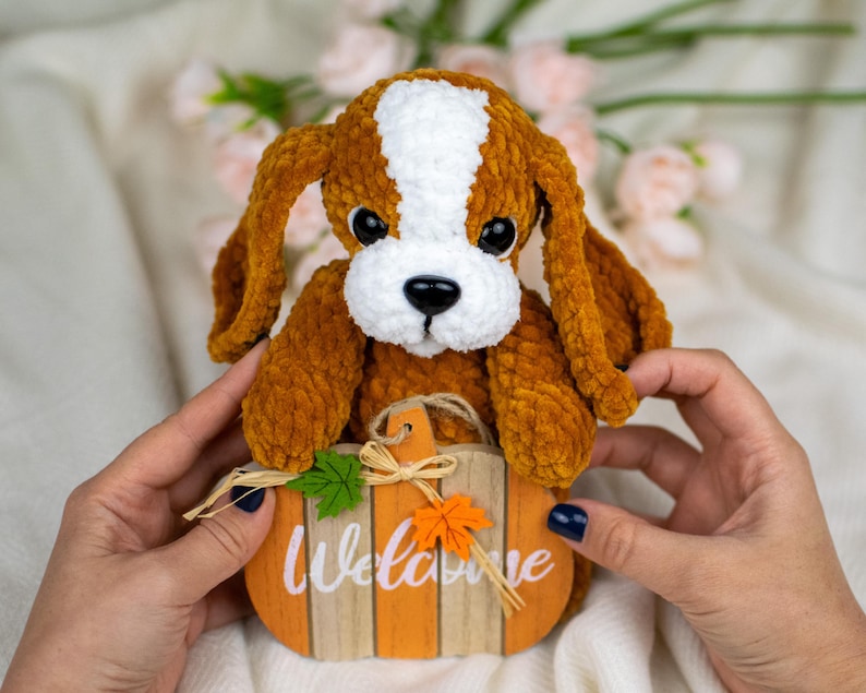May include: A plush, brown and white dog toy with large, floppy ears and black eyes. The dog is holding a small, orange pumpkin-shaped sign that reads "Welcome" in white lettering. The sign has decorative leaves and a twine bow.