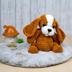 May include: A handmade, plush dog toy with brown and white fur, large floppy ears, and black eyes. The dog is sitting on a gray, fluffy surface. A small ceramic mushroom house and wooden discs are in the background.
