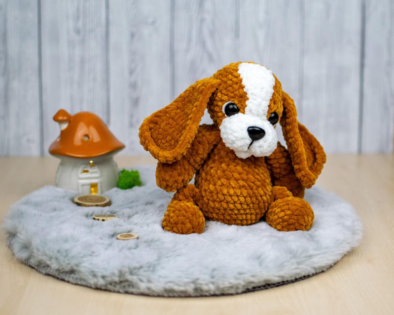 May include: A handmade, plush dog toy with brown and white fur, large floppy ears, and black eyes. The dog is sitting on a gray, fluffy surface. A small ceramic mushroom house and wooden discs are in the background.