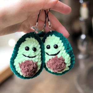 May include: Two handmade avocado-shaped charms. Each charm features a green crocheted exterior, a light green interior, and a brown crocheted pit. The avocados have black eyes and smiling mouths. They are attached to black cords.