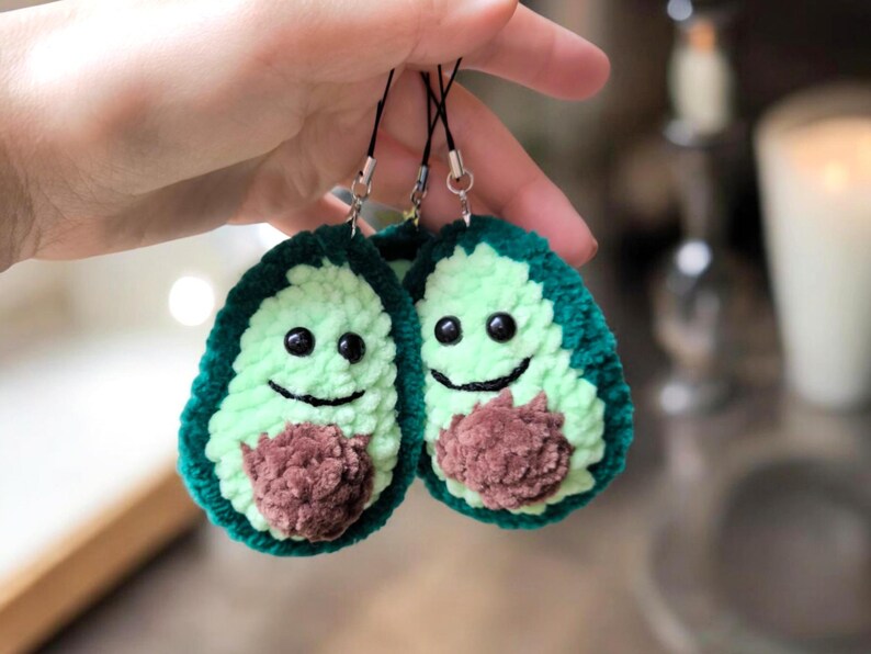 May include: Two handmade avocado-shaped charms. Each charm features a green crocheted exterior, a light green interior, and a brown crocheted pit. The avocados have black eyes and smiling mouths. They are attached to black cords.