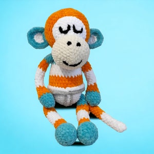 May include: A handmade crochet monkey doll with orange, white, and blue accents. The monkey has a white face, closed eyes, and a smiling mouth. The arms and legs are striped orange and white, with blue accents on the ears and feet.