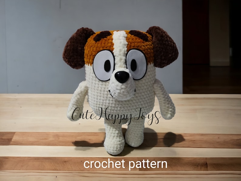 May include: Crochet pattern for a white, brown and orange dog plush toy. The toy has big eyes and a black nose. The toy is standing on a wooden surface. "Cute Happy Toys" is written on the surface.