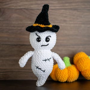 Op de afbeelding: Een handgemaakt wit gehaakt spook met een zwarte heksenhoed met een oranje band. Het spook heeft zwarte gestikte details en boze ogen. Twee oranje gehaakte pompoenen met groene stelen op de achtergrond. Halloween thema.