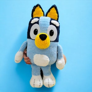 May include: A blue and yellow crocheted stuffed animal dog with big eyes and a white chest. The dog is sitting on a blue background.