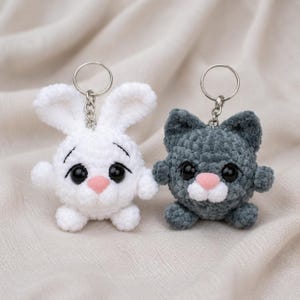 May include: Two crochet keychains featuring a white rabbit and a dark gray cat. Both plush figures have large black plastic eyes, small pink noses, and silver metal key rings attached to the top of their heads with short silver chains.