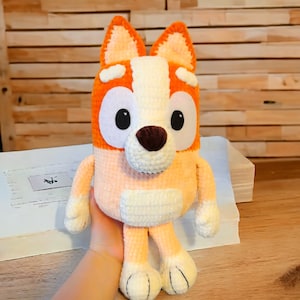 May include: A crocheted stuffed dog toy with orange, white, and brown fur. The dog has large, black eyes and a brown nose.