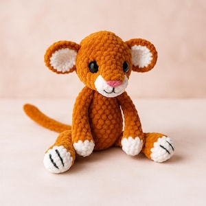 May include: A handmade, orange crochet lion cub with white accents on the ears, paws, and muzzle. The plush toy has black eyes and a pink nose. The lion cub is sitting with its legs extended and tail resting on the surface.