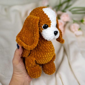 May include: A handmade plush dog toy in shades of brown and white. The dog has large, floppy ears, a white patch on its head, and black eyes. The soft, textured material gives the toy a cuddly appearance.