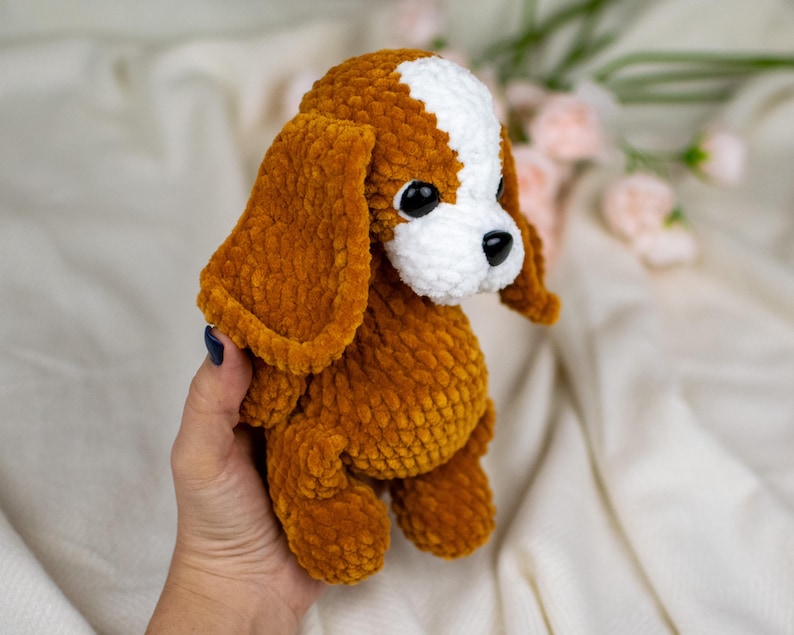 May include: A handmade plush dog toy in shades of brown and white. The dog has large, floppy ears, a white patch on its head, and black eyes. The soft, textured material gives the toy a cuddly appearance.