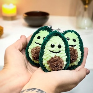May include: Three plush avocado keychains with smiling faces and brown centers. The keychains are green and light green, with black eyes and a smiling mouth. The keychains are held in a person's hands.