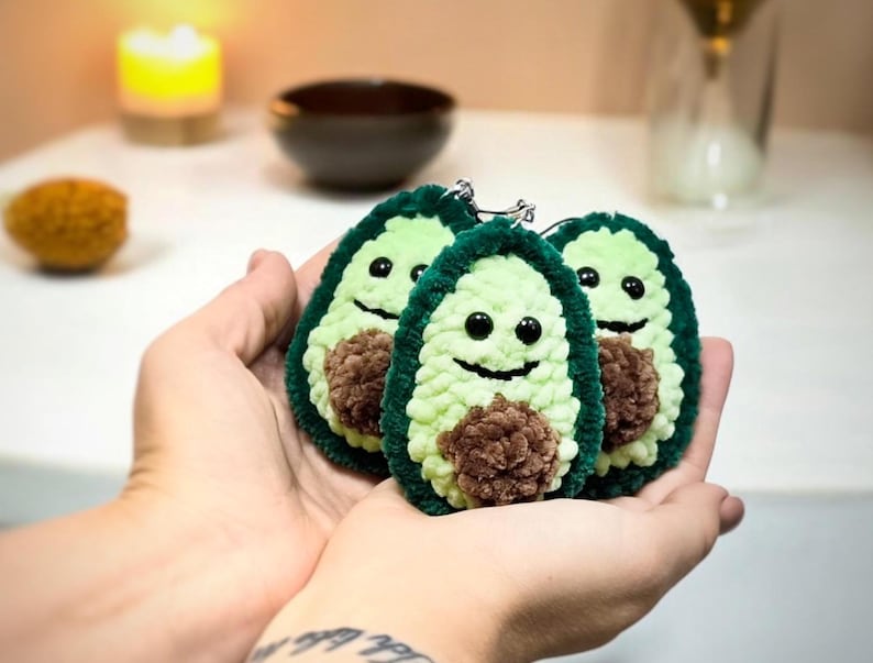 May include: Three plush avocado keychains with smiling faces and brown centers. The keychains are green and light green, with black eyes and a smiling mouth. The keychains are held in a person's hands.