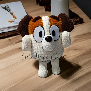 May include: A crocheted stuffed dog toy with brown and white fur, large eyes, and a black nose. The toy is standing on a wooden surface.