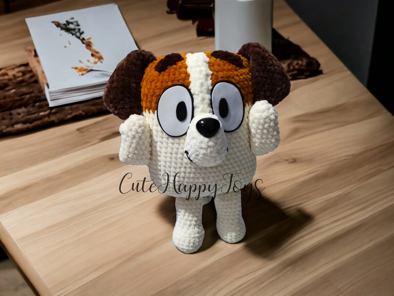 May include: A crocheted stuffed dog toy with brown and white fur, large eyes, and a black nose. The toy is standing on a wooden surface.