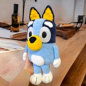 May include: A blue and yellow crocheted stuffed toy dog with white accents. The toy has large, round eyes and a black nose.