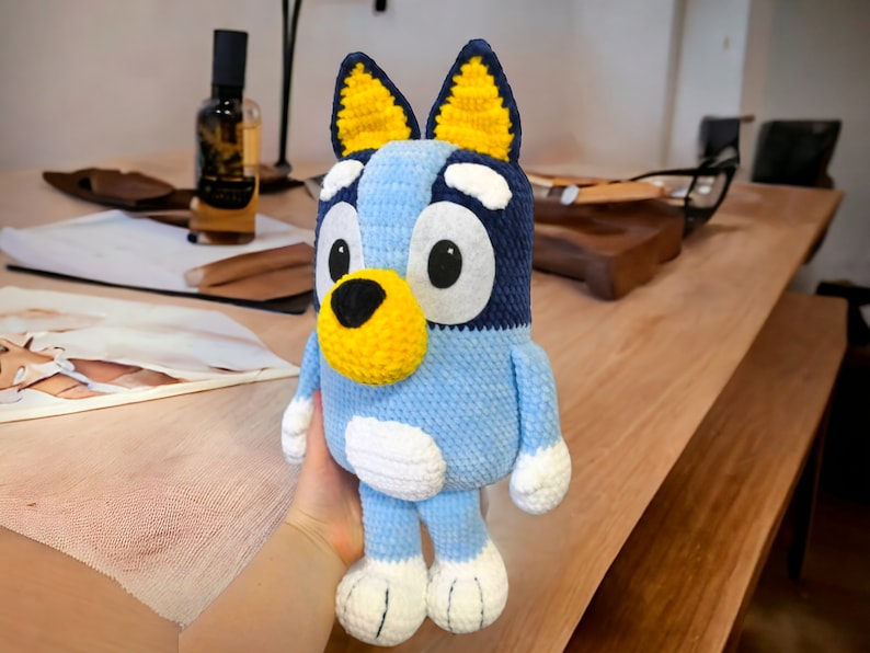 May include: A blue and yellow crocheted stuffed toy dog with white accents. The toy has large, round eyes and a black nose.