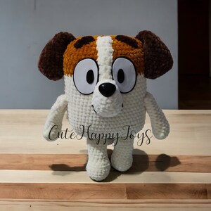May include: A crocheted plush toy dog with brown ears, white fur, and brown and white markings on its face. The toy has large, white eyes and a black nose. The toy is standing on a wooden surface.