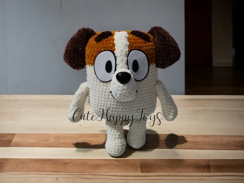 May include: A crocheted plush toy dog with brown ears, white fur, and brown and white markings on its face. The toy has large, white eyes and a black nose. The toy is standing on a wooden surface.