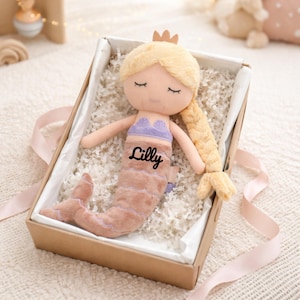 Puede incluir: Una muñeca de sirena de peluche con cabello rubio y una corona dorada, presentada en una caja de regalo con el nombre "Lilly" impreso en la cola. La muñeca tiene un top morado y una cola rosa y beige. La caja está llena de papel triturado blanco.
