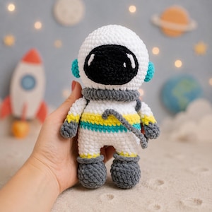 May include: A handmade white crochet astronaut doll with a black visor, gray accents, and yellow and teal stripes. The astronaut has gray boots and a gray neck piece. The doll is approximately 6 inches tall and is a space-themed toy.