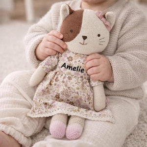 Könnte beinhalten: Handgemachte Katzenspielpuppe mit braun-cremefarbenem Kopf, trägt ein geblümtes Kleid mit dem Namen "Amelie" aufgestickt. Die Puppe hat rosa gestrickte Füße und eine rosa Schleife. Die Puppe wird gehalten.